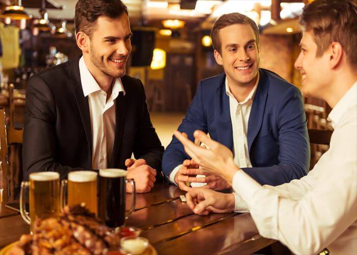 Three young businessmen in suits are smiling, talking and drinking beer while sitting in pub