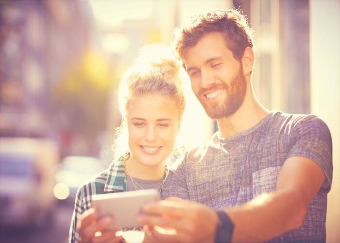 Smiling couple taking selfie through smart phone. Male and female partners spending leisure time in city. They are wearing casuals on sunny day.